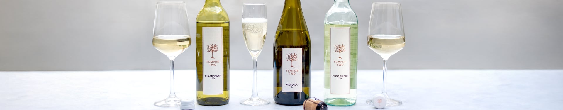 Three bottles of Tempus Two white wines—Chardonnay, Prosecco, and Pinot Grigio—are displayed alongside three matching wine glasses on a light surface, showcasing the brand's modern label design and fresh wine styles.